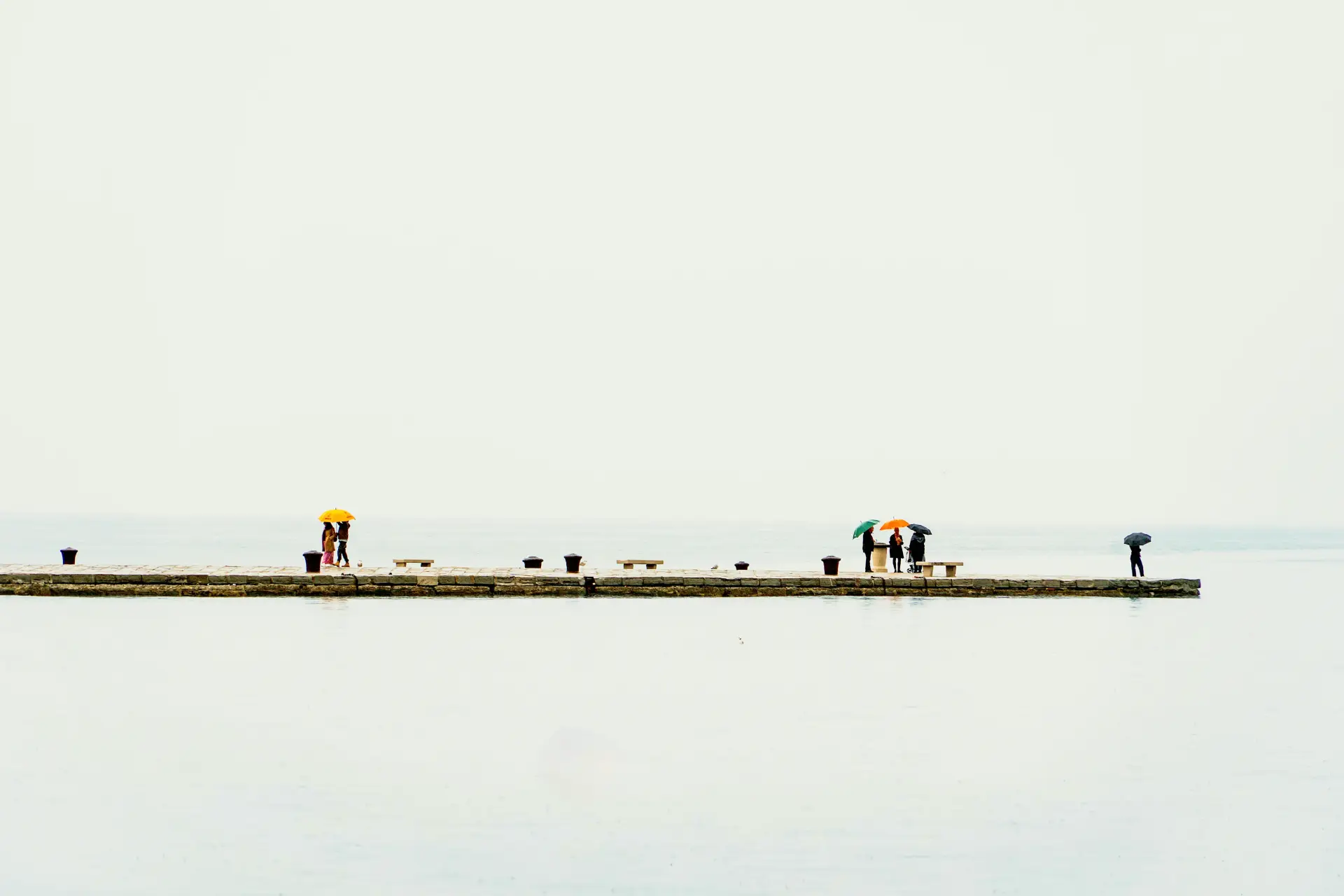 A long jetty over pale water, a few distant walkers under colourful umbrellas