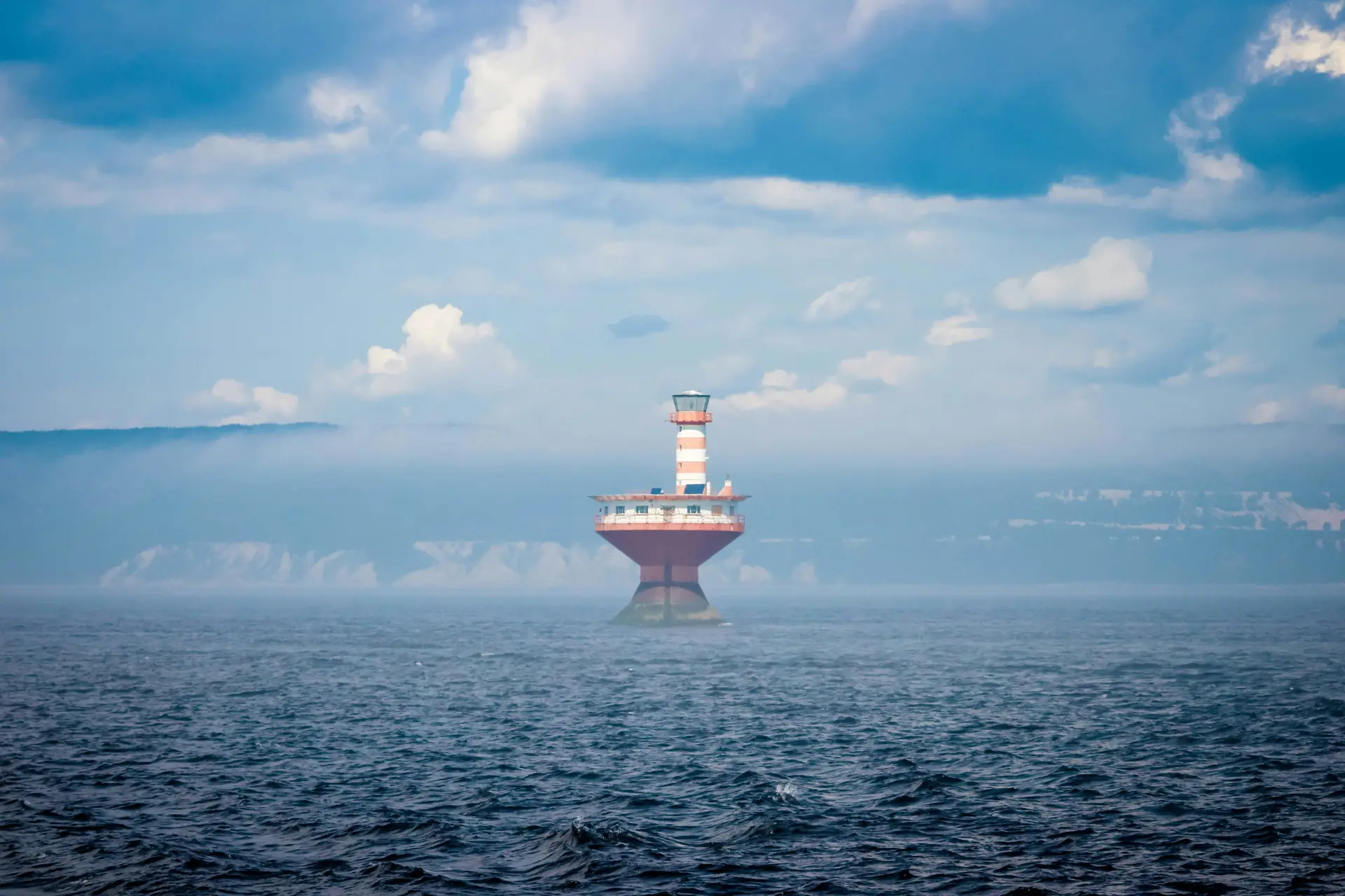 Phare rayé rouge et blanc dressé en pleine eau, au large d’une côte voilée de brume