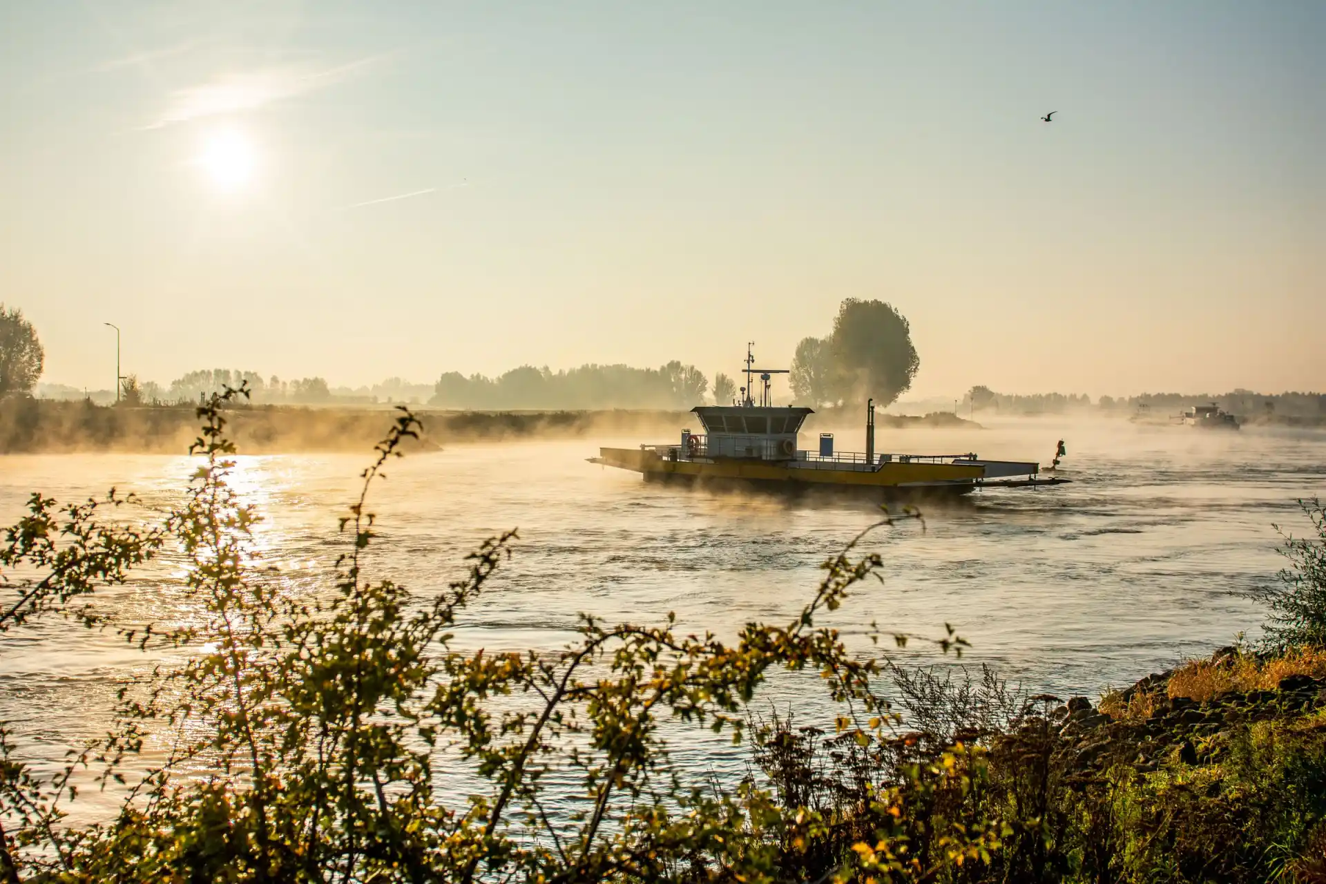 Traversier franchissant une rivière dans la brume du matin, soleil bas sur l’eau